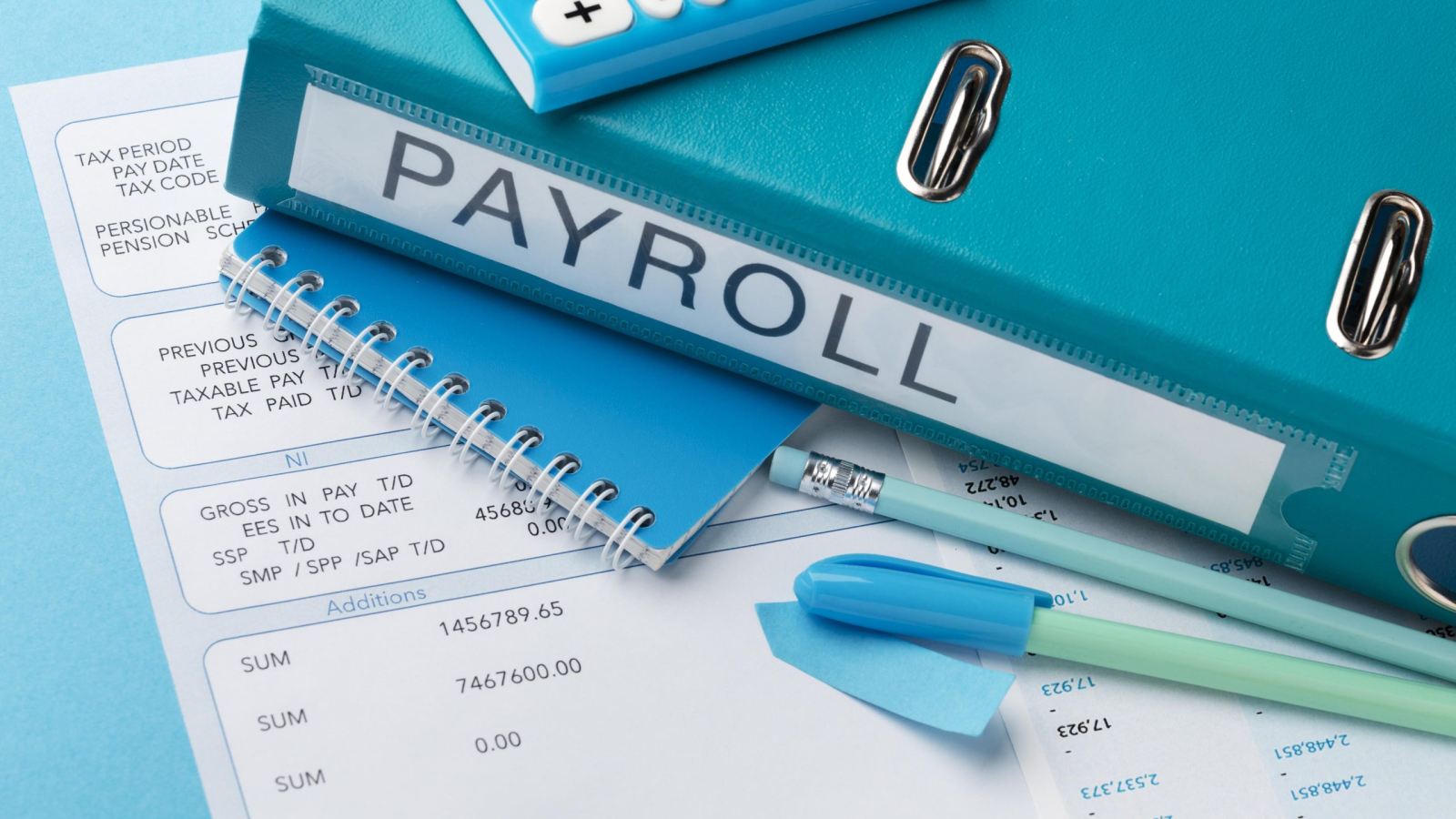 A close-up shot of a blue payroll binder with a calculator, pens, sticky notes, and payroll documents scattered on a blue desk.