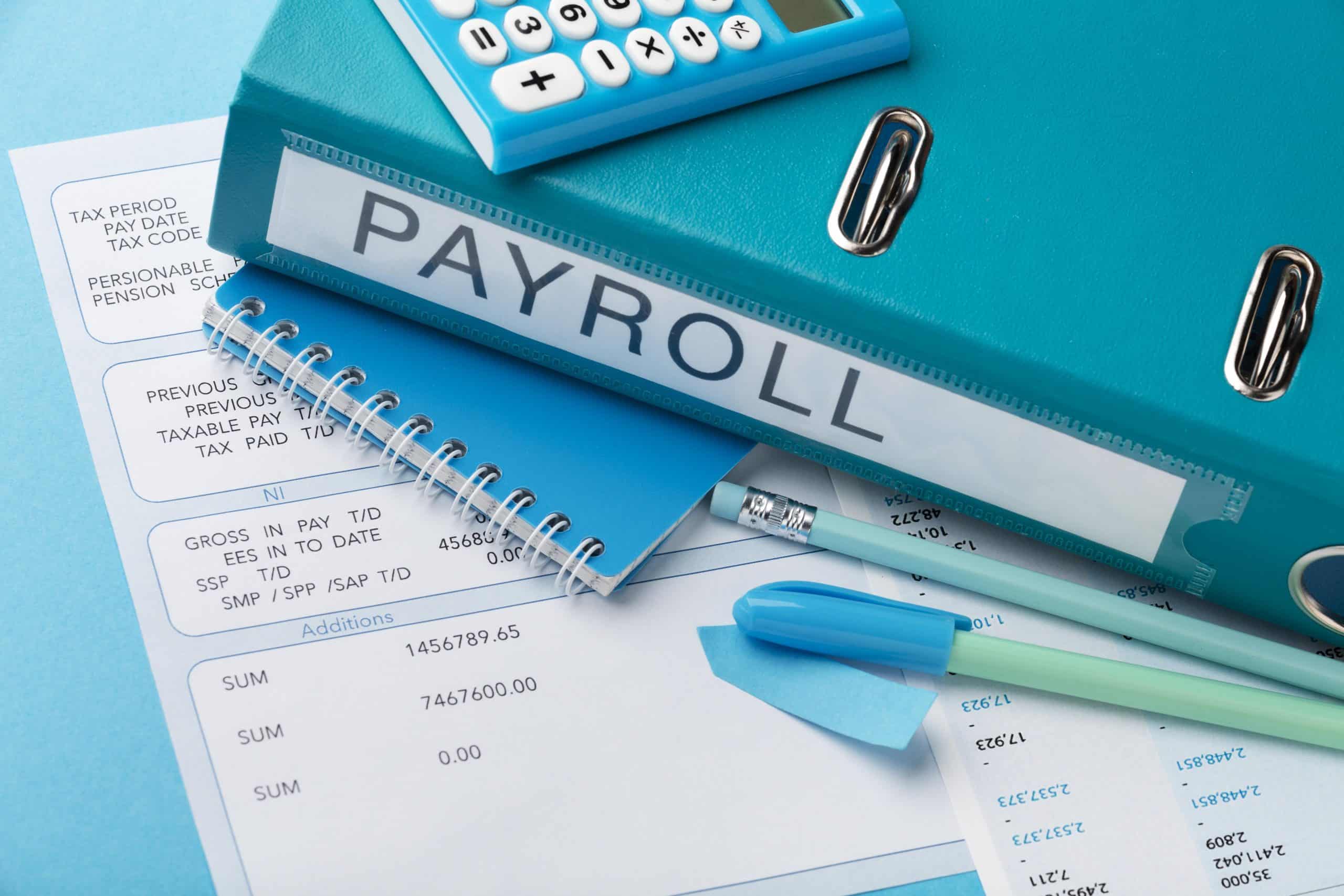 A close-up shot of a blue payroll binder with a calculator, pens, sticky notes, and payroll documents scattered on a blue desk.