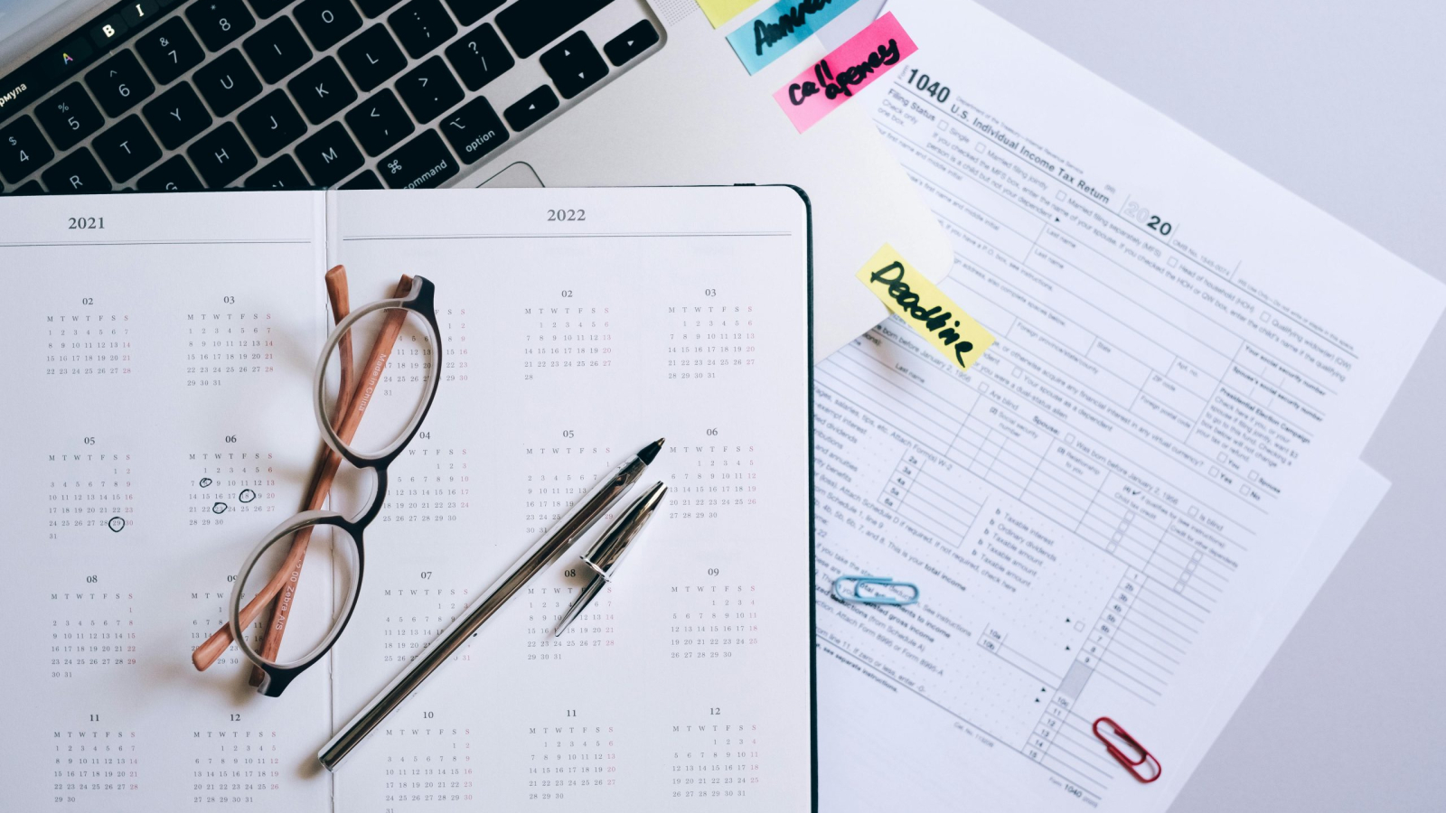 Overhead view of a desk with an open 2021/2022 planner, eyeglasses, a pen, a 1040 tax form, and sticky notes marked "Tax," "Annual," and "Deadline."