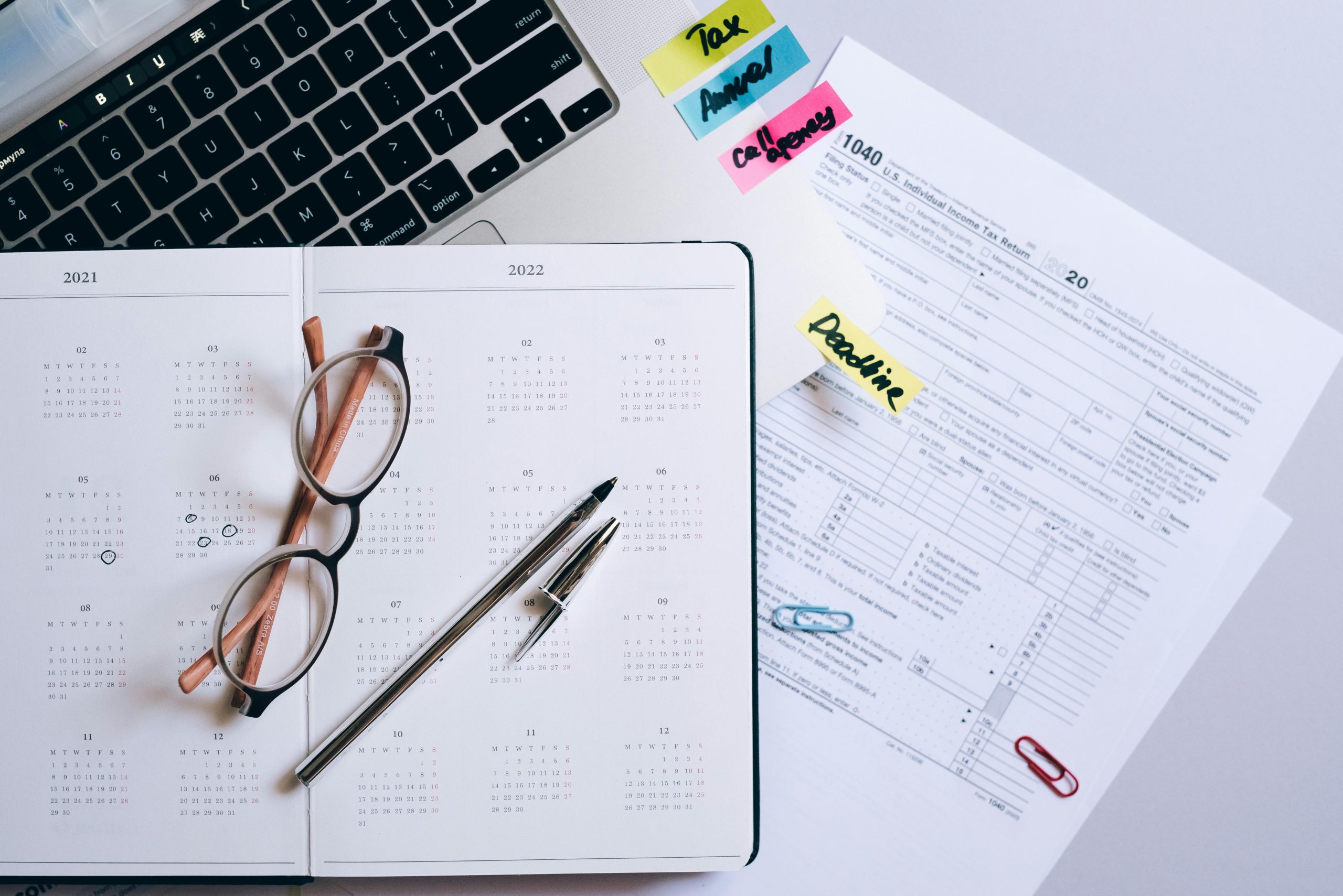 Overhead view of a desk with an open 2021/2022 planner, eyeglasses, a pen, a 1040 tax form, and sticky notes marked "Tax," "Annual," and "Deadline."