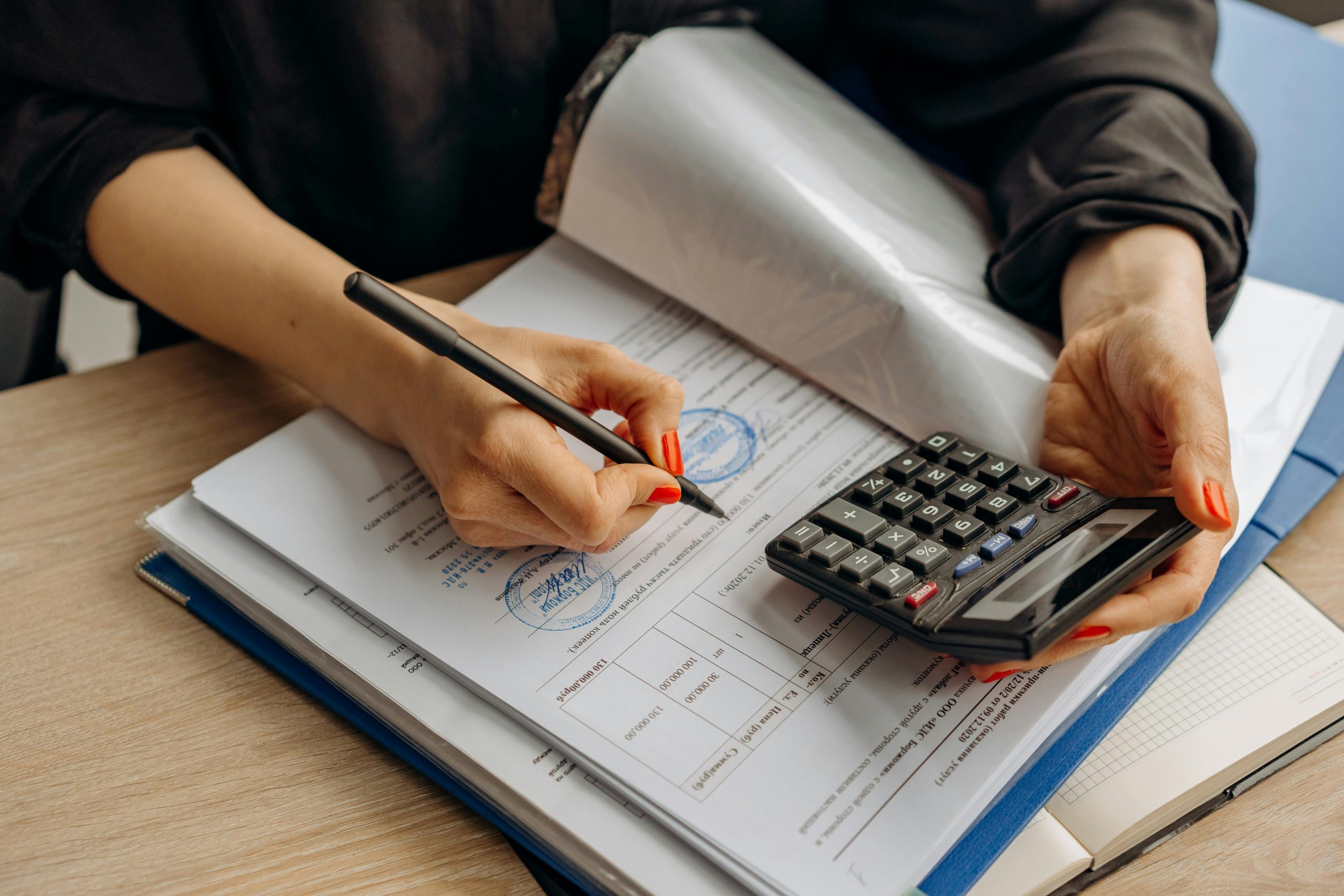 
A person with red nail polish uses a pen to sign a document stamped with a seal, while holding a calculator nearby on a wooden surface.