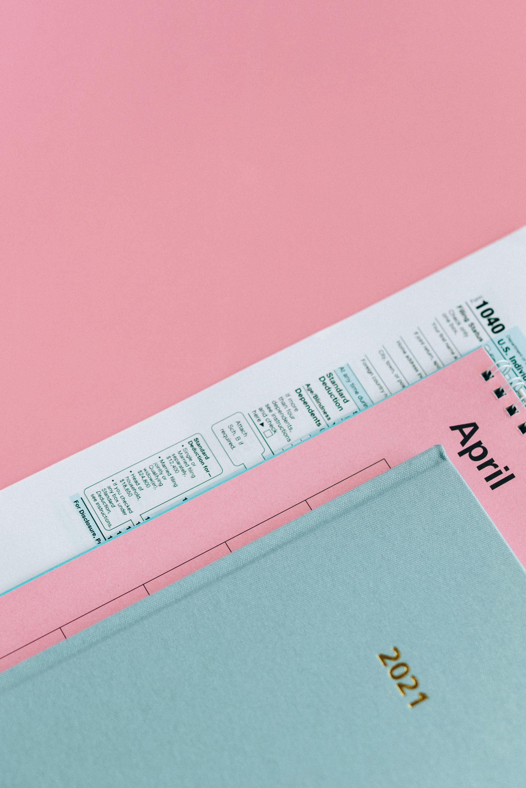 Close-up, high-angle shot of a U.S. 1040 tax form, a 2021 planner/diary, and a pink calendar page showing April, set against a pink background.