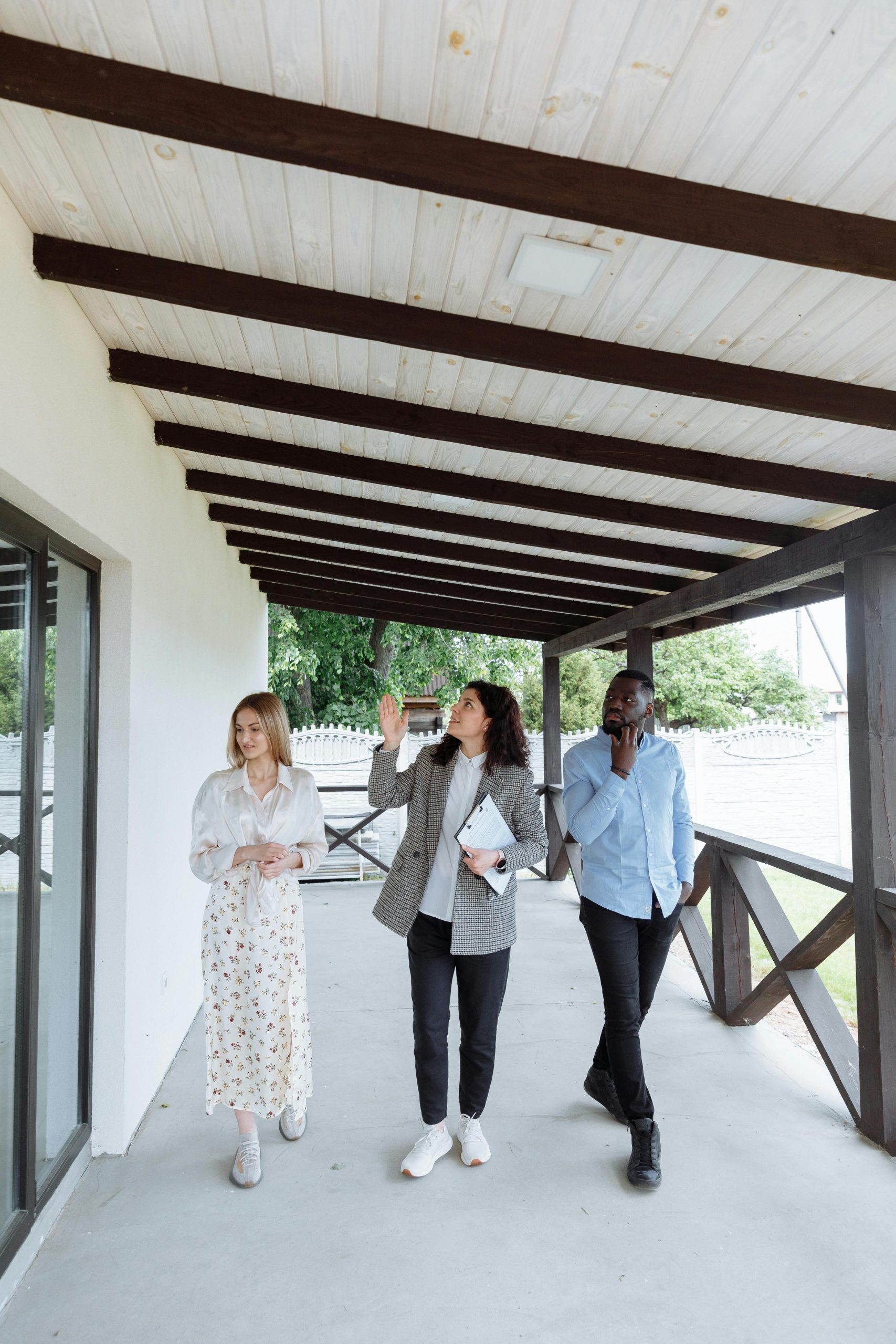 Three professionals—two women and one man—walk and discuss property on the covered porch of a modern building, possibly a real estate showing.