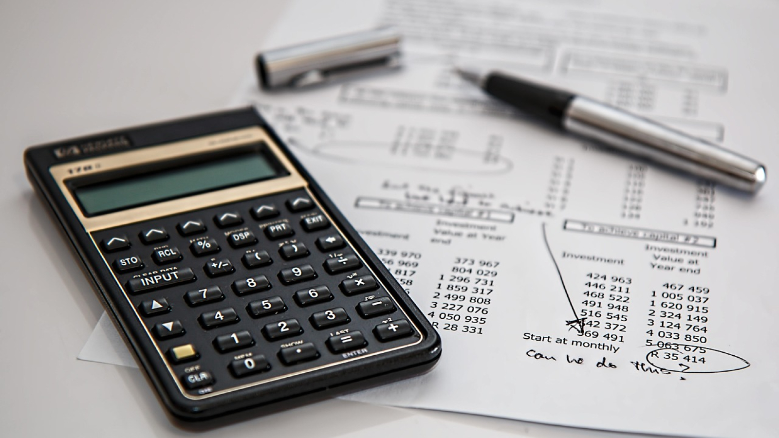 Close-up of a business desk with a black financial calculator, handwritten notes on a spreadsheet, and a chrome fountain pen.