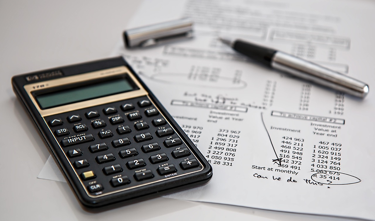 Close-up of a business desk with a black financial calculator, handwritten notes on a spreadsheet, and a chrome fountain pen.