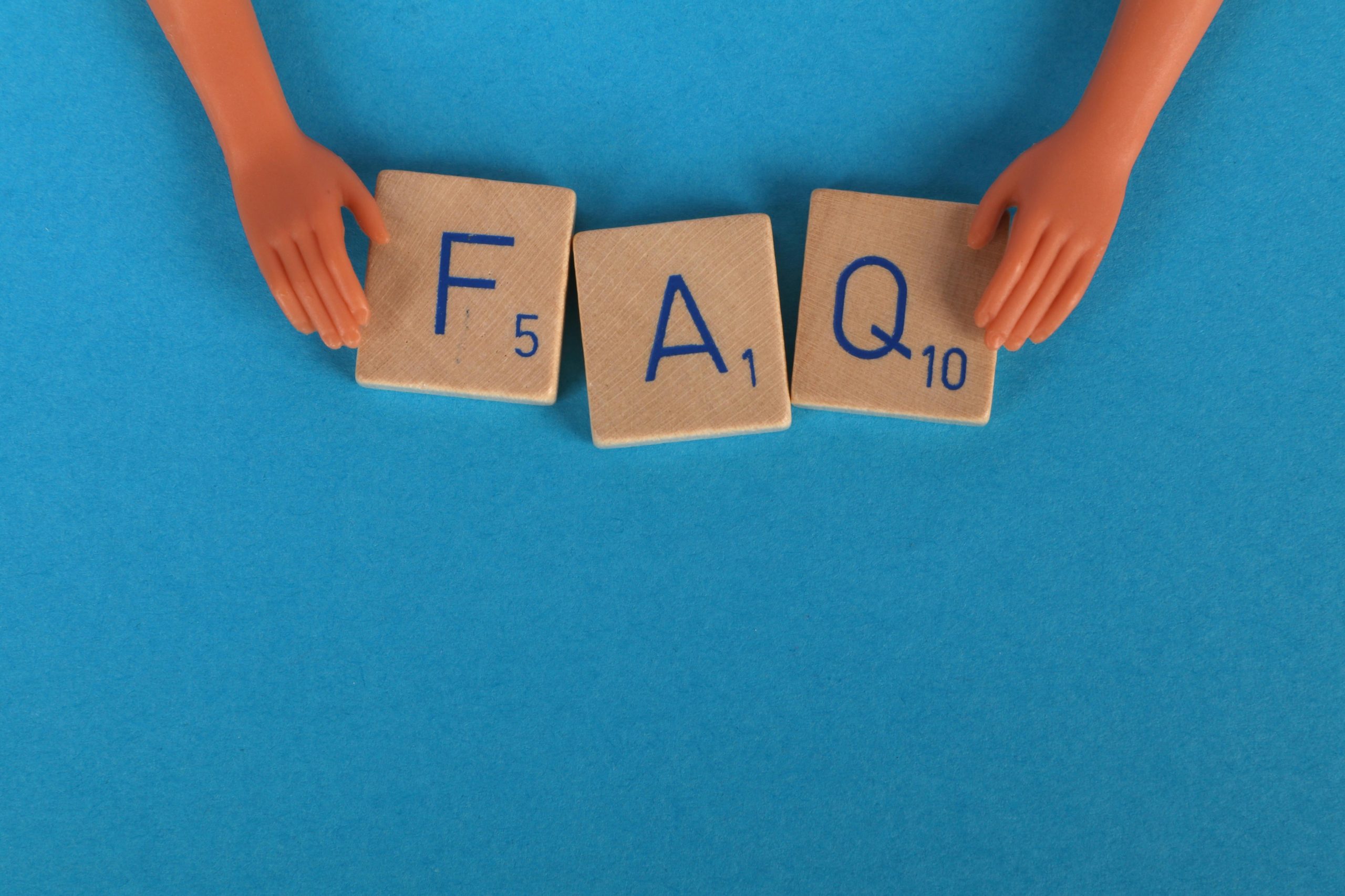 Two plastic doll hands frame three wooden letter tiles spelling out the acronym FAQ (Frequently Asked Questions) on a bright blue background.