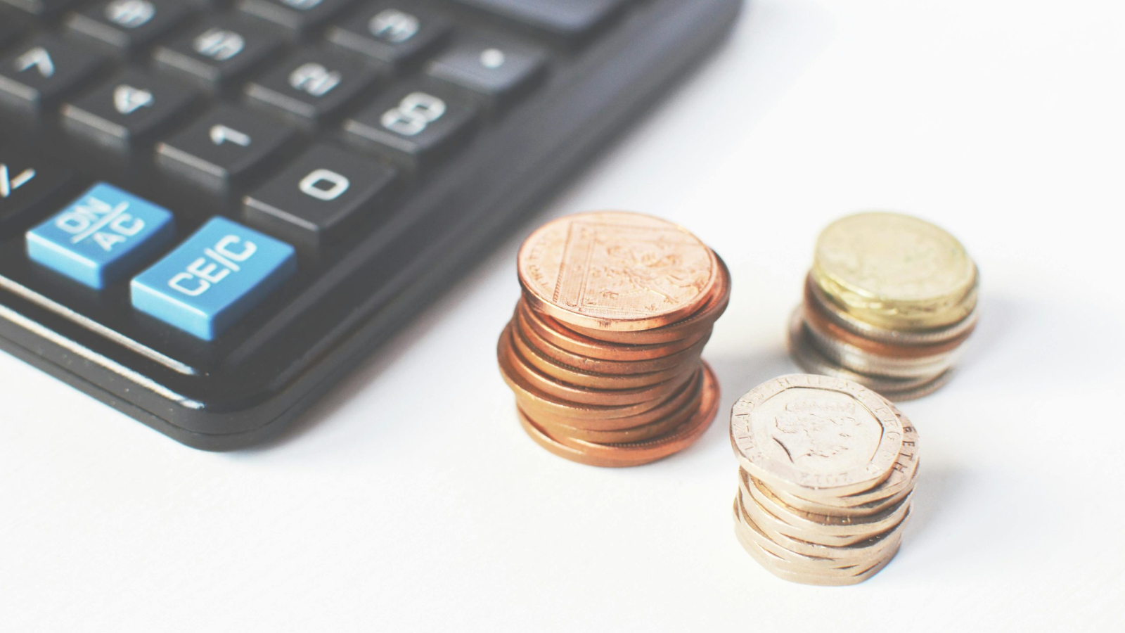 A close-up shot of several stacks of bronze and silver coins next to a dark calculator on a white surface, suggesting finance or budgeting.