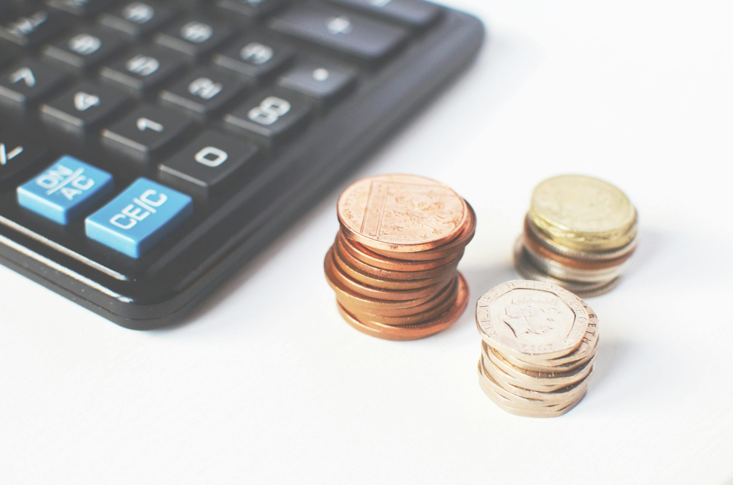 A close-up shot of several stacks of bronze and silver coins next to a dark calculator on a white surface, suggesting finance or budgeting.