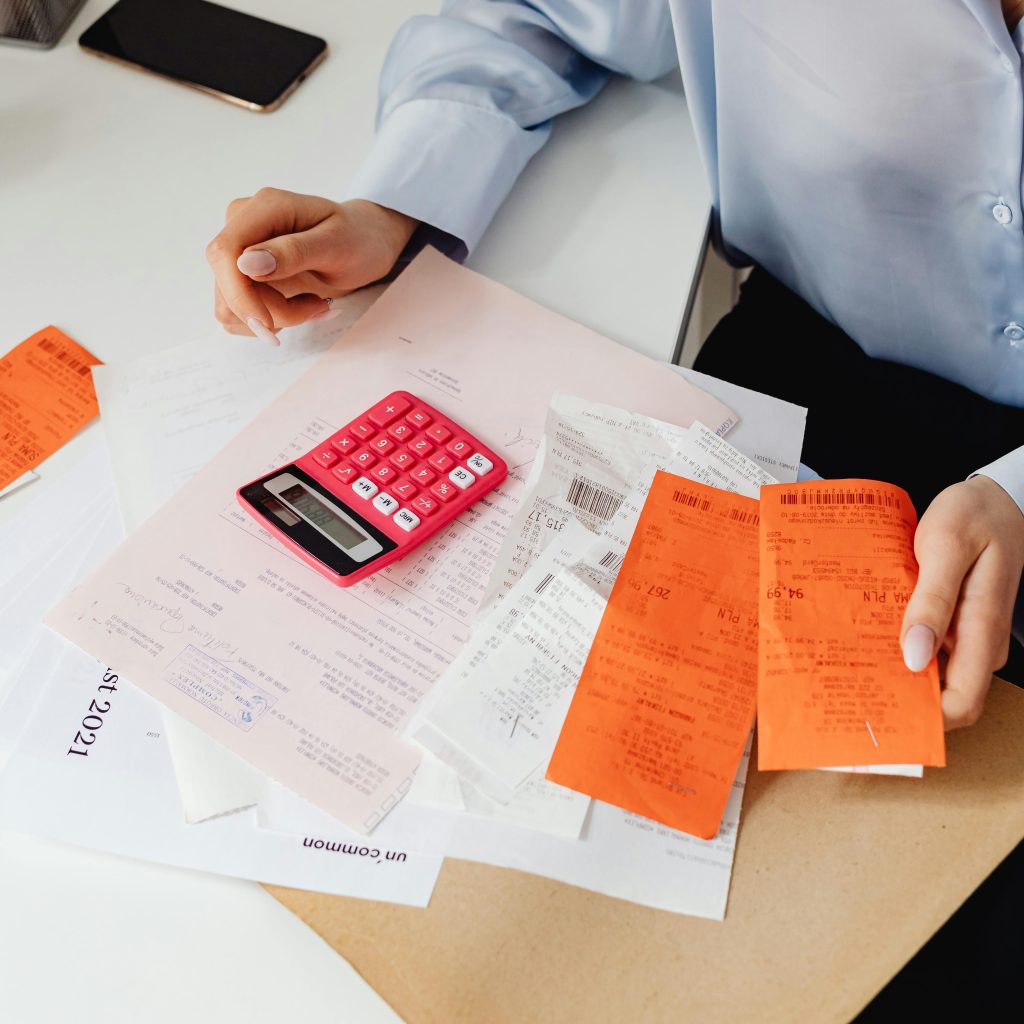 Overhead view of hands sorting a large pile of paper and bright orange receipts, with a small pink calculator in the center.