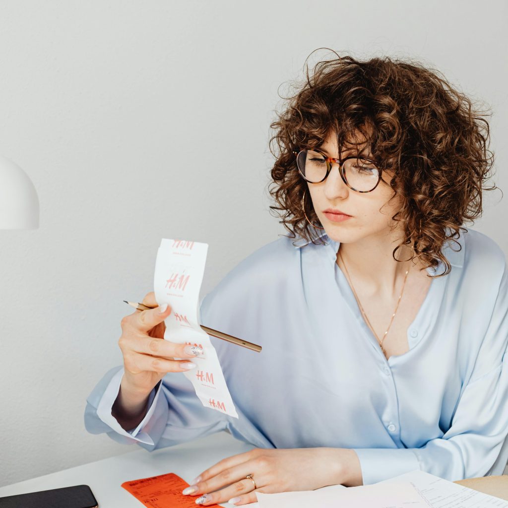A woman with curly hair and glasses reviews a long H&M receipt with a pen, while a pink calculator and orange receipts sit on her desk.