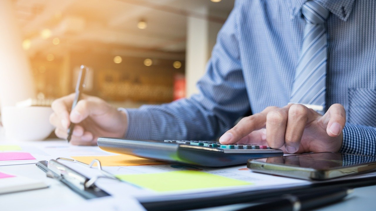 A close-up shot of an accountant in a blue shirt and striped tie using a calculator and writing with a pen on financial documents.
