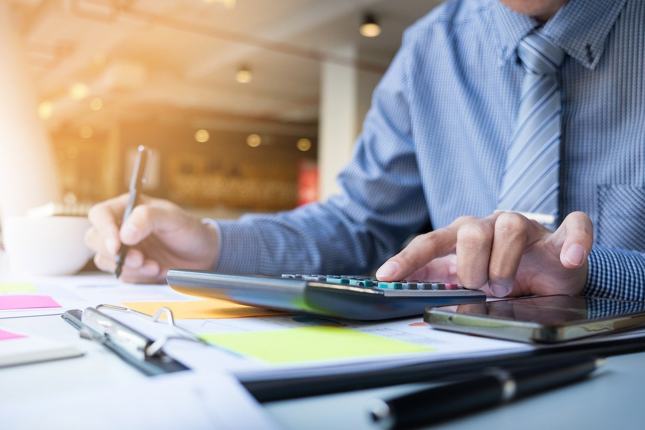 A close-up shot of an accountant in a blue shirt and striped tie using a calculator and writing with a pen on financial documents.