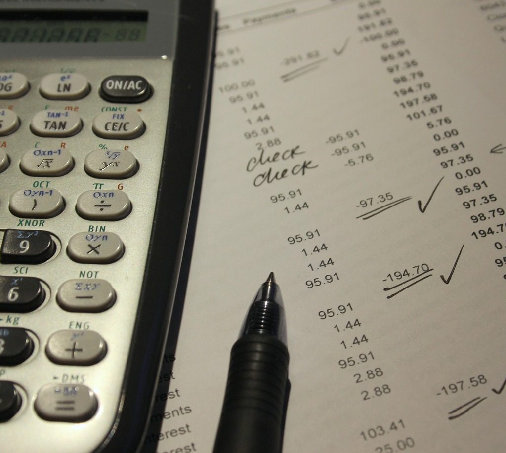 A close-up of a scientific calculator, a black pen, and a document with a column of numbers, calculations, and check marks.