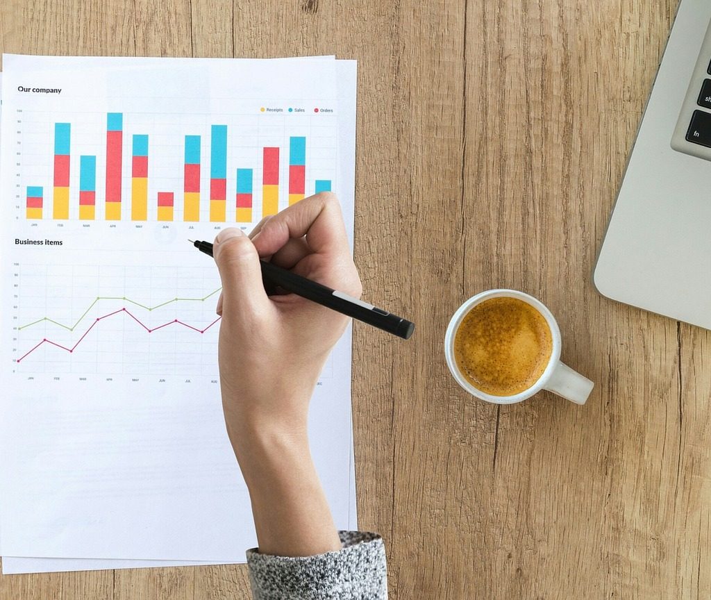 A man in a blue dress shirt and striped tie uses a calculator and writes notes on a document on a sunny desk, focusing on finance.