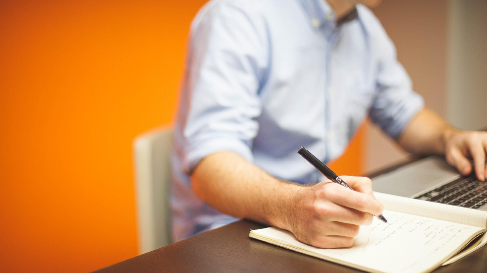 Man in light blue shirt working at a dark desk, writing in a notebook with a black pen while using a laptop. Bright orange wall in background.