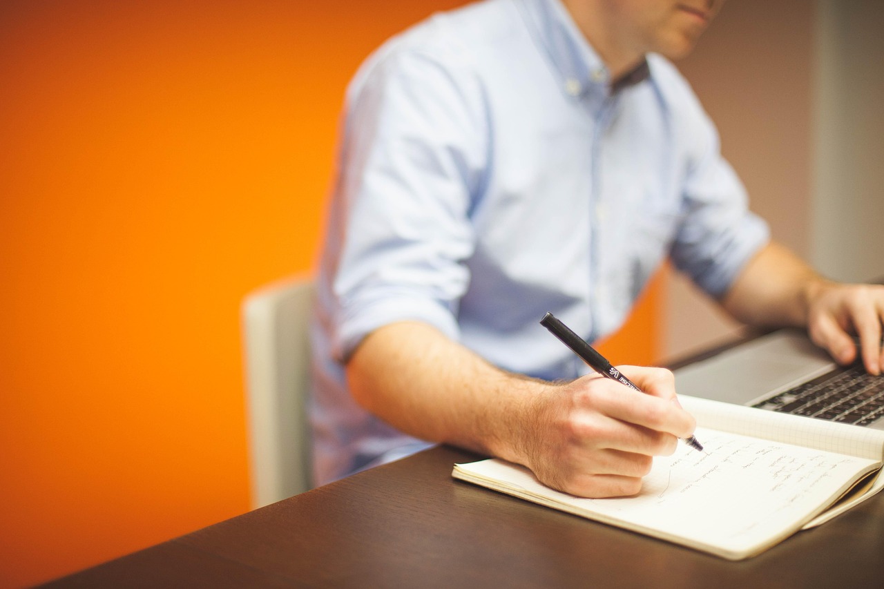 Man in light blue shirt working at a dark desk, writing in a notebook with a black pen while using a laptop. Bright orange wall in background.